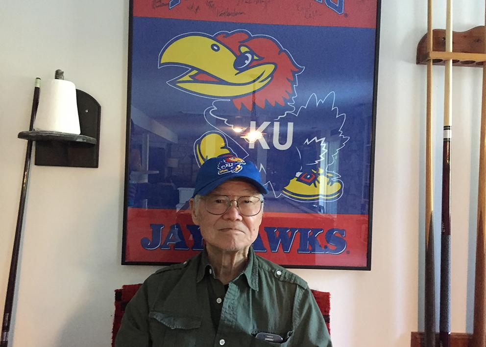 Ted Kuwana sitting in front of Jayhawk poster